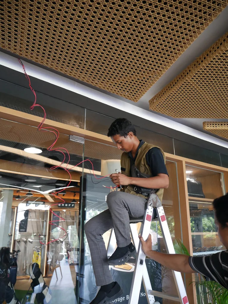 Technician on a stepladder pulling cable beside a glass partition under a rattan ceiling